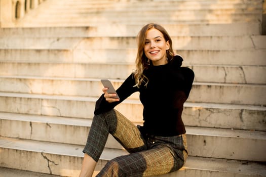 Trendy young woman with mobile phone sitting at stairs outdoor
