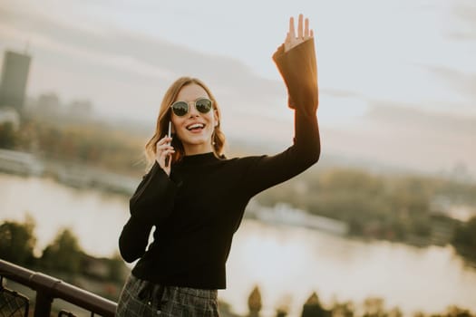 Portrait of young woman holding mobile phone while standing on walkway by the river