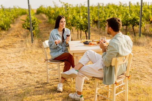Couple relaxes at a rustic table in a sunlit vineyard, savoring wine and gourmet snacks. They share laughter and joy, surrounded by lush vines and the warmth of the evening