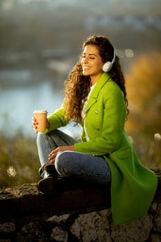 Pretty young woman listening music with smartphone and takeaway coffee by the river at sunny autumn day