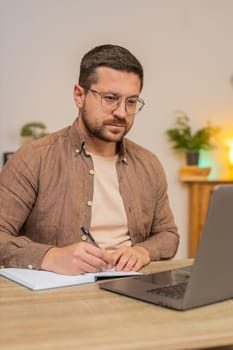 Young businessman writing down or taking down notes while attending online video class or office meeting on laptop computer at home office desk. Guy freelancer support services looking at webcam.
