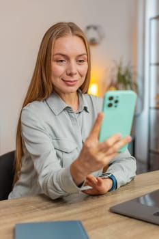 Happy mature Caucasian woman sitting at table desk using mobile phone smiling at home apartment. Girl freelancer texting share messages on smartphone social media applications online watching movie.
