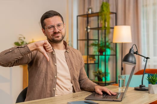 Dislike. Upset unhappy man using laptop and showing thumbs down sign gesture, expressing discontent, disapproval dissatisfied bad work at home office. Displeased young guy freelancer at table desk.