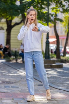 Young woman worrying to be punctual, with anxiety checking time on watch, running late to transport, being in delay, deadline outdoors. Girl teenager tourist walking in city street. Town lifestyles.