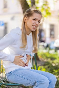 Sick ill young woman suffering from period cramps painful stomach ache outdoors. Girl teenager holding belly feeling abdominal menstrual pain sitting on urban city street. Abdominal pain gastritis.