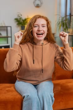 Happy young woman watching TV shouting, celebrating success, winning, goal achievement good news, lottery luck, victory sitting on sofa in living room. Adult girl showing double thumbs up at home.