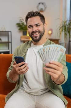 Successful rich young Caucasian man holding and waving dollar bill money fan sitting on sofa couch at home. Happy adult guy winning online lottery game planning vacation calculating budget concept.