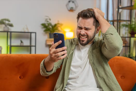Upset displeased Caucasian man using smartphone sitting on sofa couch in living room. Young adult guy in casual clothes touching head loses becoming surprised by sudden lottery results or bad news.