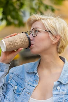 Happy Caucasian young blonde woman enjoying morning coffee hot drink and smiling outdoors. Relaxing, taking a break. Girl walking on urban city center street, drinking coffee to go. Town lifestyles.