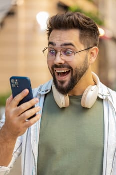 Excited amazed Caucasian man outside using mobile phone device winning online shocked male tourist in city win smartphone bet celebrate victory achievement success. Guy standing on urban city street.