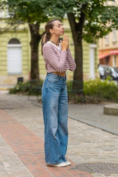 Portrait of young pretty woman praying with closed eyes to God asking for blessing, help, health, forgiveness outdoors. Girl clasping hands wishing luck in urban city street. Town religion lifestyles