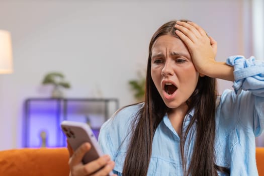 Sad displeased Caucasian woman using smartphone typing browsing, loses becoming surprised sudden lottery game results, bad news, fortune loss fail, deadline, virus. Young girl at home room on couch