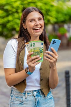 Happy young woman counting money euro cash, using smartphone calculator app, satisfied of income for planned vacation gifts. Lovely girl standing in urban city sunny street. Town lifestyles outdoors