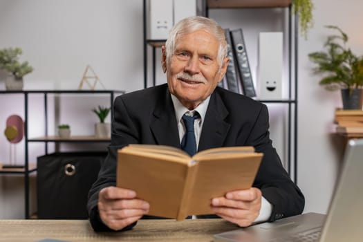 Bored Caucasian senior businessman reading book, literature, taking a rest after work. Grandfather freelancer guy in suit with laptop relaxing with head in hand at home office table. Business people.