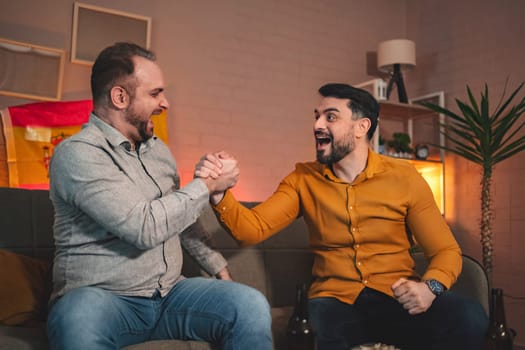 In a warm and inviting living room, two friends are engaged in a lively arm wrestling match, their expressions full of joy and competitive spirit. A Spanish flag adds to the festive atmosphere.