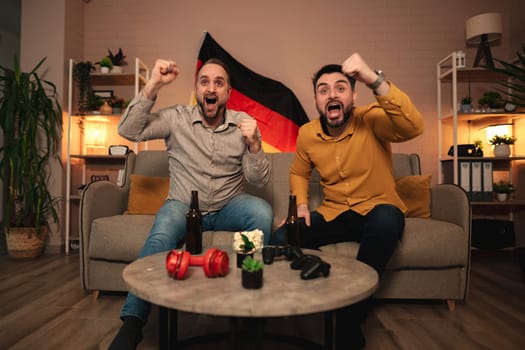 Two friends cheer joyfully from a comfortable couch, celebrating a hard-fought victory. The room is warmly lit, featuring a German flag, drinks, and playful decorations.