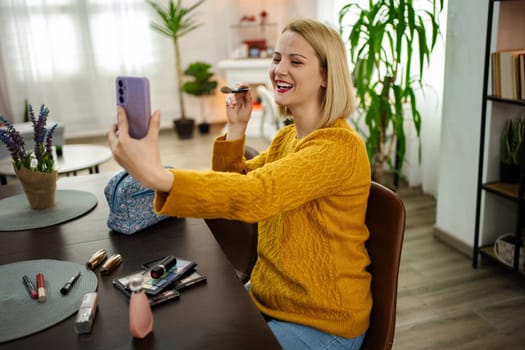 A cheerful woman is engaged in a lively video call, applying her makeup with a mirror in a warmly decorated living space filled with plants. Sunlight streams in, creating a bright atmosphere.