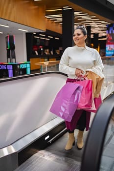 A cheerful young woman ascends an escalator with bright shopping bags.