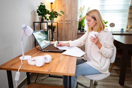 Woman enjoying tea while working on a laptop in a bright room.