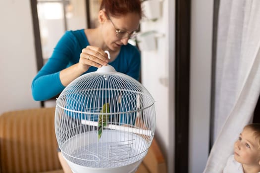 Mother closing a white round birdcage with a green parrot inside, her child looking at the bird