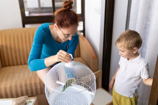 Mother closing parrot cage while her son watches in bright living room