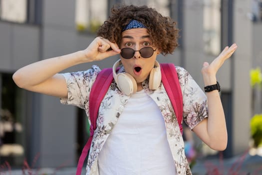 Oh my God, Wow. Happy young man taking off sunglasses looking surprised at camera with big open eyes, shocked by sudden victory, game winning, lottery goal achievement, good news outdoors city street.