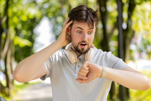 Happy active man runner jogging in city park and looking at smartwatch. Excited male using fitness tracker and checking result celebrating success. Athlete guy is satisfied with results of training.