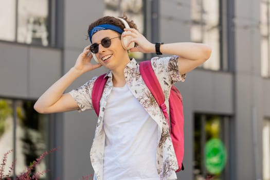 Happy relaxed young Caucasian man in wireless headphones dancing, listening favorite energetic disco music outdoors. Smiling overjoyed tourist guy with curly hair and backpack walking in city street.