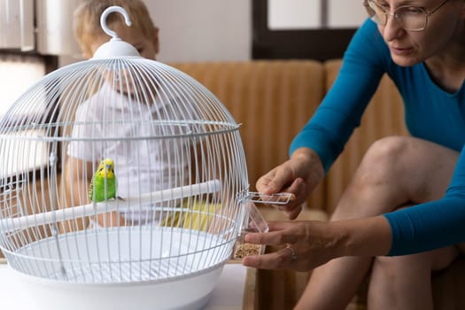 Woman carefully refills bird feeder in a cage with a green and yellow parakeet, as a child watches