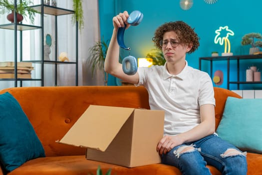 Unhappy young Caucasian man guy unpacking carton box, feeling dissatisfied with getting damaged wireless headphones order, writing negative feedback in mobile application, internet shopping concept.
