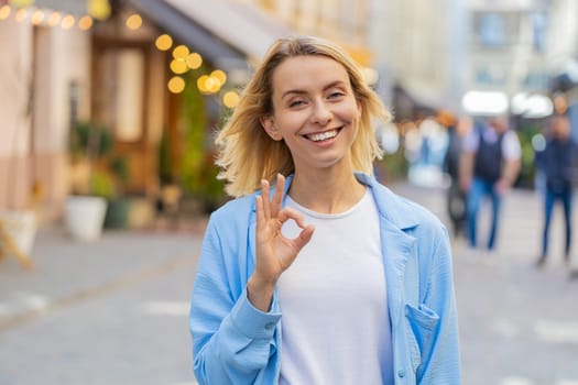Okay. Happy cheerful young Caucasian woman looking approvingly at camera showing OK gesture positive like sign, approve something good celebrate win. Joyful lady standing in urban city street outdoors