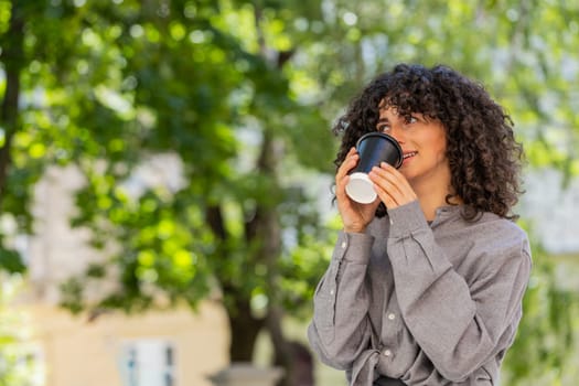 Happy Caucasian young woman enjoying morning coffee hot drink and smiling outdoors. Relaxing, taking a break. Lady girl walking on urban city street drinking coffee to go. Town lifestyles. Sunny park
