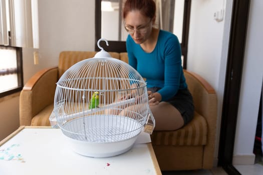 Woman changing bird food in a white cage with a green budgie parrot perched on a wooden stick, inside a house