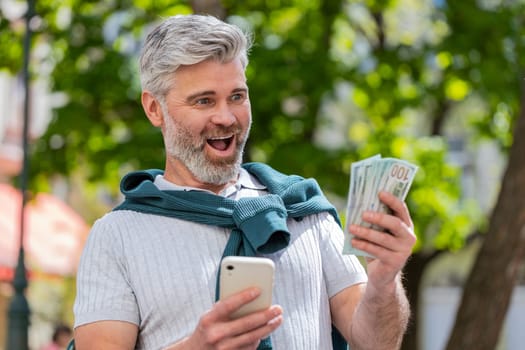 Happy rich mature man counting holding money dollar cash, use smartphone calculator app, plans to order gifts and food delivery online, booking hotel room. Bearded guy tourist in city street, outdoors