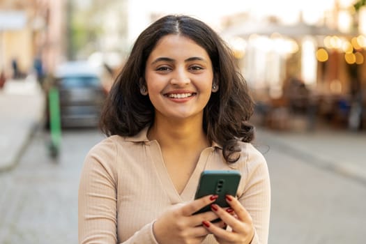 Happy smiling Indian young woman using smartphone typing text messages browsing internet, finishing work, looking at camera outdoors. Hispanic girl walking on urban city street. Town lifestyles