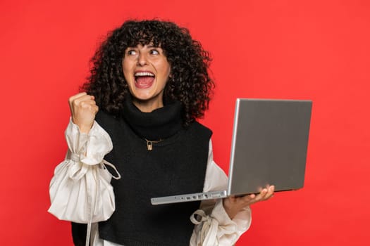 Happy excited Caucasian woman typing on laptop, working on project, enjoying results, winning lottery game celebrating success, online shopping victory good email news. Pretty girl on red background