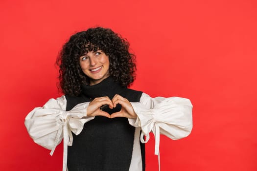 Woman in love. Smiling attractive Caucasian young woman with curly hair makes heart gesture demonstrates love sign expresses good positive feelings and sympathy. Girl isolated on red background