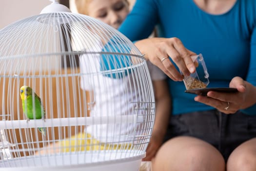 Mom pouring birdseed into a small dish for a parakeet while her daughter watches