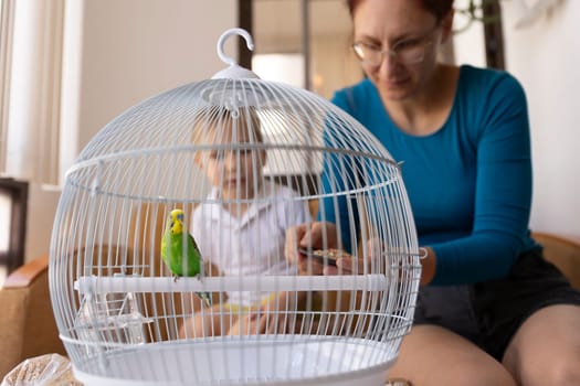 Mother and child feeding a green budgie perched inside its cage, promoting family bonding and responsible pet ownership