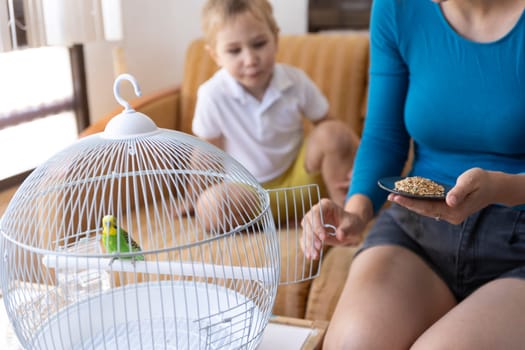 Mom opening birdcage door, holding plate with bird food, and her son watching a green budgie