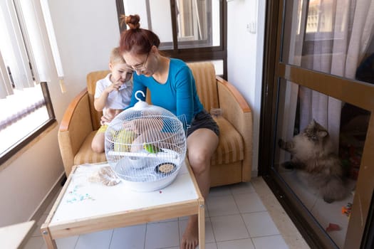Mother and son sitting on sofa, interacting with pet parakeet in cage, while cat curiously observes from behind balcony door