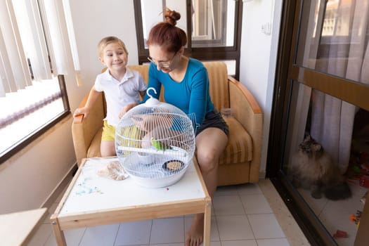 Mother and son interacting with their pet budgie in a cage on the balcony, while a cat observes from behind a glass door