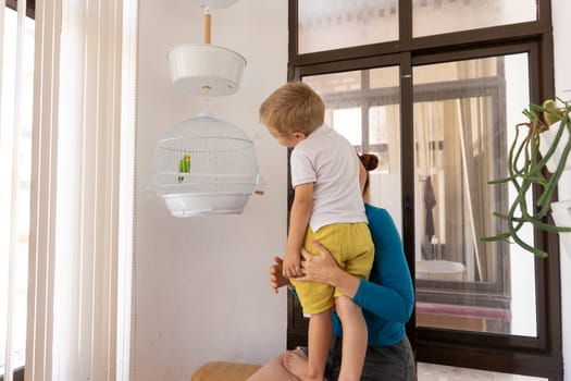 Mother holding her son so he can look at a green parrot in its cage hanging from the ceiling