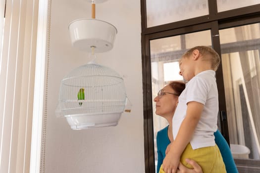 Mother and son admiring a green parrot in a cage hanging from the ceiling