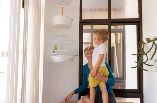 Smiling mother lifting her toddler son to see a parakeet in a white cage hanging on the balcony