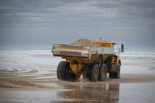 A dump truck carries loads of sand along the biscarosse dune coastline. The vehicle works to combat erosion as it traverses the wet, reflective sands near the ocean.