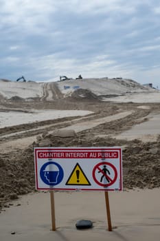 Signs indicate restricted access due to construction efforts aimed at managing coastal erosion while heavy machinery operates on the sand at Biscarosse beach under a cloudy sky.