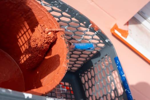 A paint roller rests in a bucket filled with reddish-brown paint. Surrounding tools and a work environment indicate ongoing home improvement activities in good weather.