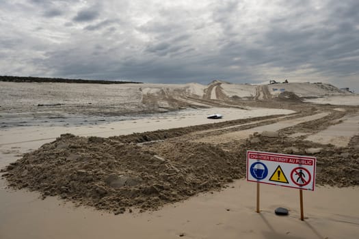 Signs indicate restricted access due to construction efforts aimed at managing coastal erosion while heavy machinery operates on the sand at Biscarosse beach under a cloudy sky.