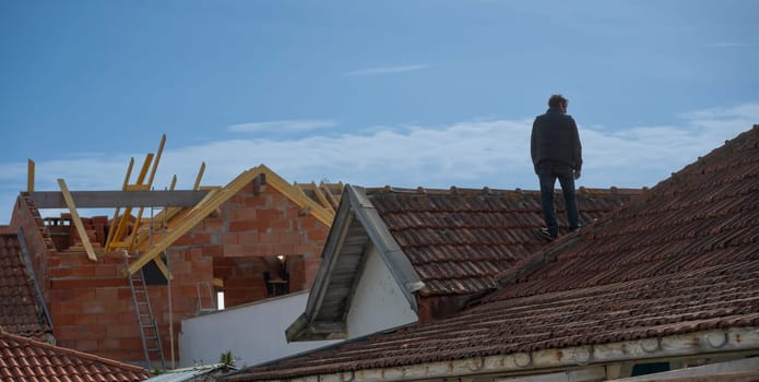 A man is positioned on the sloped roof of a house, which is undergoing construction. Exposed wooden beams indicate ongoing work, while a ladder leans against the structure.
